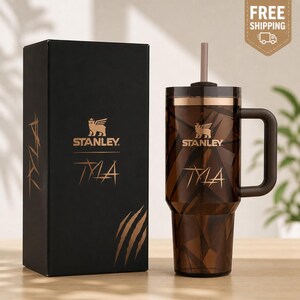 May include: A brown and copper Stanley TLA tumbler with a matching box. The tumbler has a geometric pattern and a handle. The box is black with gold text and a logo. The tumbler includes a straw.