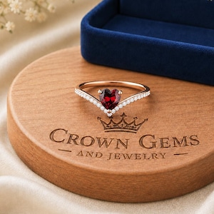 Pode incluir: Um anel de ouro rosa com uma pedra vermelha em forma de coração no centro de uma banda em V decorada com pequenas pedras brancas. O anel repousa sobre uma base circular de madeira gravada com CROWN GEMS AND JEWELRY, perto de uma caixa azul.