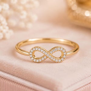 May include: A gold ring featuring a horizontal infinity symbol centrepiece. The infinity loop is encrusted with small, clear, round-cut stones that catch the light, set against a polished gold band that completes the circular shape of the ring.