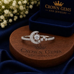May include: A silver-toned ring featuring a crescent moon motif encrusted with small clear stones and a single round brilliant-cut center stone. The band is accented with a series of small star-shaped settings, each containing a tiny clear stone.