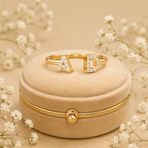 Puede incluir: Un anillo abierto dorado con las letras A y D, cada una incrustada con pequeñas piedras transparentes. El anillo está colocado sobre un recipiente redondo de textura aterciopelada beige con una banda de metal dorado en el centro y un cierre circular.