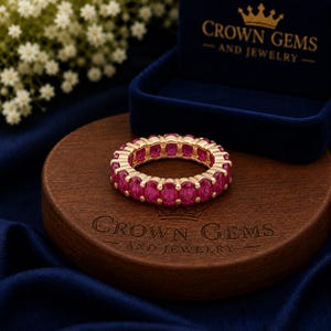 Peut inclure: Une bague alliance éternité composée d'une rangée continue de pierres précieuses ovales rose vif serties sur un anneau en métal doré. La bague est posée sur un socle en bois circulaire gravé du texte Crown Gems and Jewelry.