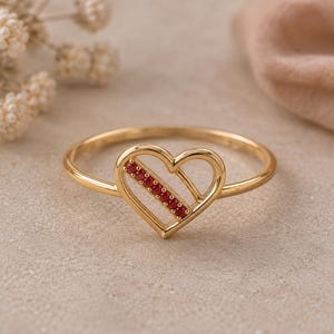 Pode incluir: Um anel de ouro com um elemento central em forma de coração. O contorno do coração é feito de ouro polido, com uma secção diagonal cravejada com cinco pequenas pedras preciosas redondas de um vermelho vibrante. O aro é fino, liso e circular, mantendo um tom dourado uniforme.