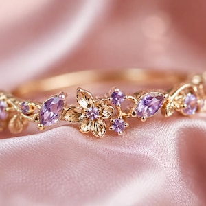 May include: A gold ring featuring a central flower motif with engraved petals and small purple gemstones. The band is adorned with marquise-cut and round purple stones arranged in a vine-like pattern, creating a delicate and detailed botanical design.