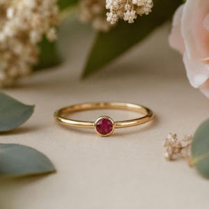 May include: A gold ring with a small, round, ruby gemstone set in the center. The ring is thin and delicate, with a polished finish. The background features soft, blurred flowers and foliage.