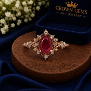 Può includere: Un anello in oro rosa con un rubino rosso intenso taglio ovale al centro, circondato da un alone a forma di stella di piccoli diamanti bianchi. L'anello è appoggiato su una base in legno con l'incisione CROWN GEMS AND JEWELRY.