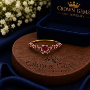 May include: A gold ring featuring a central heart-shaped red gemstone flanked by smaller round red stones and a small gold bow detail. The ring is displayed on a wooden base engraved with the text CROWN GEMS AND JEWELRY near a blue box.