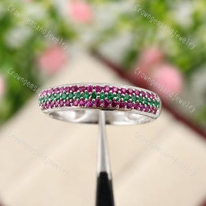 Pode incluir: Um anel de prata com uma faixa de fileiras alternadas de pequenas pedras preciosas redondas vermelhas e verdes. O anel é segurado por uma ferramenta de metal contra um fundo desfocado. O anel é uma joia.