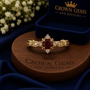 May include: A gold ring featuring a central round deep red gemstone surrounded by smaller clear stones in a star-shaped setting. The band has an intricate metalwork design. The ring is placed on a wooden base with the text CROWN GEMS AND JEWELRY.