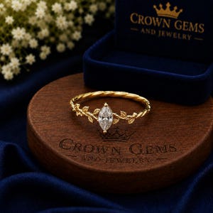 May include: A gold ring featuring a marquise-cut clear gemstone at the center, flanked by delicate leaf-shaped metal details on a twisted band. The ring rests on a circular wooden base engraved with the text CROWN GEMS AND JEWELRY.