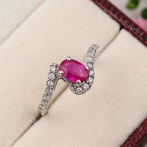 Könnte beinhalten: Ein silberner Ring mit einem großen ovalen Rubin im Zentrum. Das Band des Rings umschließt den Rubin, mit kleinen, runden, klaren Edelsteinen entlang der Kurve. Der Ring wird in einer Schmuckschatulle präsentiert.