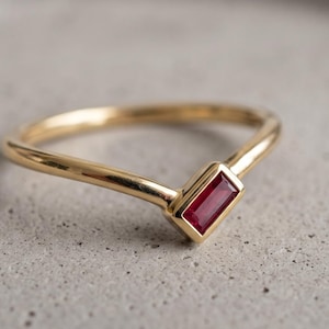 May include: A gold ring featuring a V-shaped band and a single rectangular baguette-cut ruby gemstone set in a bezel setting. The polished gold band curves to a point where the vibrant red stone is securely mounted in a horizontal orientation.