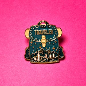 May include: A teal enamel pin shaped like a backpack with gold metal outlines. The design features the word TRAVELER in gold letters, a central buckle, and a landscape illustration of mountains, pine trees, and stars against a dark teal background.