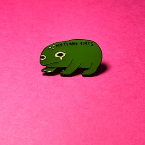 May include: A green enamel pin shaped like a frog with a sad expression and a tear under one eye. The text MY TUMMY HURTS is printed in black capital letters on the frog's back. The frog has a dark green body with lighter green spots.