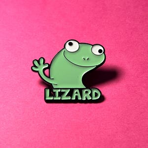 May include: A light green enamel pin featuring a cartoon lizard with large white eyes and a black outline. The lizard is waving one hand, and the word LIZARD is printed in bold, light green capital letters on a black rectangular base below.