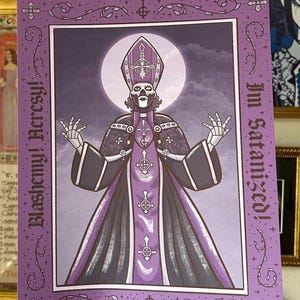 May include: A rectangular art print featuring a central figure dressed in purple and black ecclesiastical robes with a matching mitre. The figure has skeletal hands and a skull-like face. The text reads Blasphemy! Heresy! and I'm Satanized!