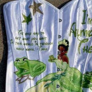 May include: A white satin corset featuring a green glitter graphic of Tiana and frogs. The text reads: The only way to get what you want in this world is through hard work. - Tiana. It also includes green and gold glitter star accents.