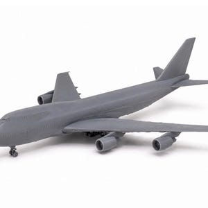 May include: A grey model of a Boeing 747 airliner. The model aircraft has four engines, wings, and a tail. The landing gear is visible. The plane is set against a white background.