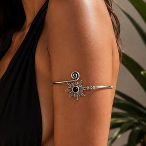 May include: A silver-toned metal arm cuff featuring a sun-shaped centerpiece with a small black circular stone at its core. The band has a spiral design on one end and a straight, textured arm that wraps around the upper arm for a secure fit.