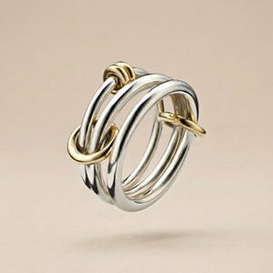 May include: A silver ring with three bands, accented with gold-coloured details. The ring features a unique design with interlocking loops and a polished finish, suitable for everyday wear or special occasions.