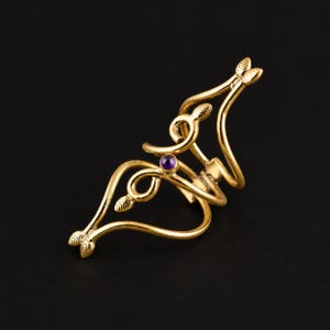 May include: A gold-toned metal ring featuring an elongated, openwork design with curved lines and small leaf-shaped accents at the terminals. A single, small, round, deep purple gemstone is set centrally within the intricate, swirling metal structure.
