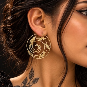 Può includere: Un grande orecchino circolare color oro caratterizzato da un design a spirale con piccoli accenti testurizzati a forma di pipistrello integrati lungo il filo. L'orecchino è indossato al lobo e presenta una finitura metallica lucida.