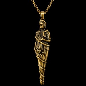 Può includere: Una collana con ciondolo in metallo color ottone raffigurante una figura scolpita con capelli intrecciati, che indossa un abito drappeggiato ed è avvolta da un serpente. La figura tiene un piccolo oggetto tra le mani ed è appesa a una catena.