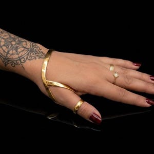 Könnte beinhalten: Ein goldfarbenes Handarmband mit einem glatten, geschwungenen Metallband, das das Handgelenk umschließt und in einen Ring übergeht, der den Daumen umfasst. Das Schmuckstück hat eine polierte, reflektierende Oberfläche und ein strukturelles Design.