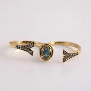 Può includere: Un anello aperto color oro con una pietra preziosa blu di forma ovale al centro, incastonata in una cornice testurizzata. Le due estremità dell'anello si dividono in forme a Y, decorate con piccole pietre chiare e scintillanti.