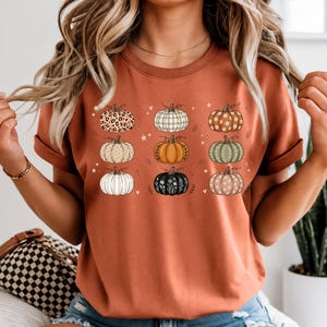 May include: A rust-colored short-sleeve t-shirt featuring a graphic print of nine pumpkins arranged in a grid. Each pumpkin displays a unique pattern, including leopard print, plaid, floral designs, and polka dots, accented by small stars and hearts.