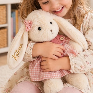 May include: A cream-colored plush rabbit toy with long ears, featuring a pink gingham dress and a small pink bow on one ear. The name Katrina is embroidered in gold thread on the rabbit's ear. The toy has dark eyes and a stitched nose.