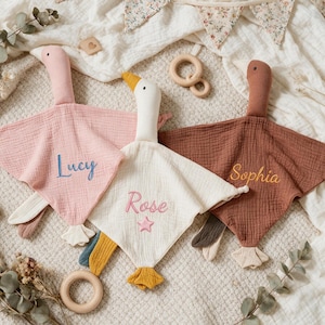 May include: Three soft muslin security blankets shaped like geese in pink, cream, and brown. The pink blanket features the embroidered name Lucy, the cream blanket features the name Rose with a star, and the brown blanket features the name Sophia.