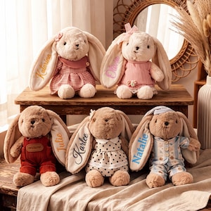 Pode incluir: Cinco coelhos de pelúcia com orelhas longas, cada um vestindo roupas diferentes, incluindo vestidos, jardineiras e pijamas. As orelhas são bordadas com os nomes Katarina, Olivia, Julie, Karlee e Natalie em várias cores de linha.