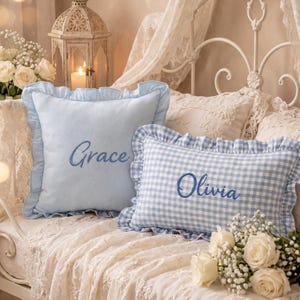 Puede incluir: Dos cojines decorativos con bordes de volantes. Un cojín cuadrado de color azul claro presenta el nombre Grace bordado en letra azul. Un cojín rectangular de cuadros vichy azul y blanco presenta el nombre Olivia bordado en letra azul.