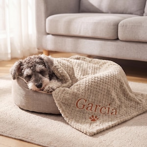 May include: A small gray and white dog sleeping in a round, light gray pet bed. The dog is covered by a textured, beige, waffle-knit blanket embroidered with the name Garcia in brown thread, accompanied by a small brown paw print icon.