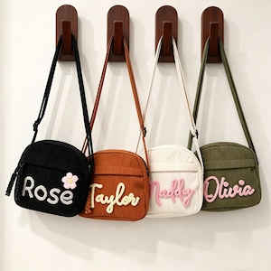 May include: Four corduroy crossbody bags hanging on wooden hooks. The bags are black, rust orange, cream, and olive green. Each bag features a personalized name in raised, textured lettering and a small crocheted flower on the black bag.