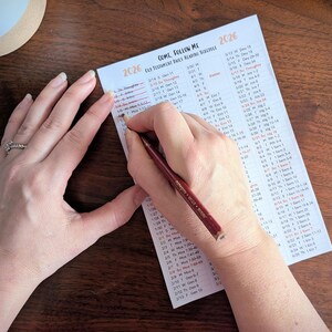 Könnte beinhalten: Hände schreiben mit einem roten Bleistift auf weißem Papier. Das Papier ist ein täglicher Leseplan für 2026 mit dem Titel "Come, Follow Me". Das Papier liegt auf einer dunklen Holzoberfläche.