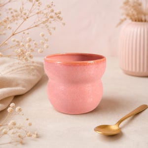 Peut inclure: Une tasse en céramique rose de forme originale, avec un bord corail. Une cuillère dorée est posée à côté. L'arrière-plan comprend un vase et des fleurs séchées, créant une esthétique douce et neutre.