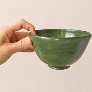 Puede incluir: Un cuenco de cerámica pequeño y redondo con un esmalte de color verde vibrante. El cuenco presenta una textura ligeramente irregular y artesanal con variaciones sutiles en el color verde y un borde oscuro definido en la parte superior, sobre una base.