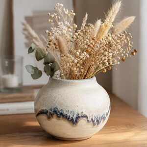 Può includere: Un vaso in ceramica rotondo con una finitura testurizzata color bianco sporco e un motivo a smalto gocciolante blu scuro attorno al centro. Il vaso è riempito con una varietà di steli botanici essiccati, tra cui erba della pampa, piccoli fiori bianchi e foglie di eucalipto.