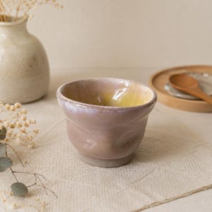 Peut inclure: Un petit bol en céramique arrondi avec un extérieur rose mauve brillant et un intérieur jaune pâle lisse. Le bol présente une forme organique légèrement irrégulière avec un bord épais et une base mate non émaillée, révélant un aspect artisanal.