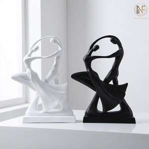 May include: Two abstract sculptures of dancers, one white and one black. Each sculpture shows two figures in a flowing pose, suggesting movement and unity. The sculptures are on a white surface. The sculptures are likely made of resin or a similar material.