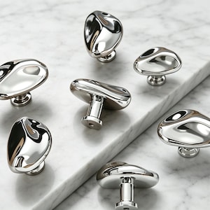 May include: Seven polished chrome cabinet knobs with organic shapes and reflective surfaces. Each knob is mounted on a short stem. The knobs are arranged on a white marble surface, reflecting the light and creating a modern aesthetic.