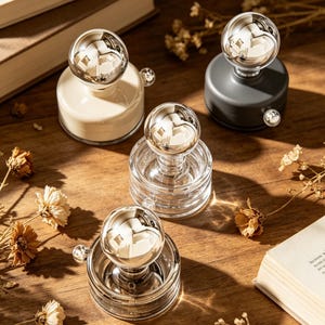 May include: A collection of decorative paperweights with a spherical, mirrored top. The bases are in various colors, including cream, clear, and dark gray. The paperweights are arranged on a wooden surface with dried flowers and a book.