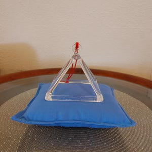 May include: A clear crystal pyramid suspended by a red string, resting on a small blue cushion. The cushion sits on a textured, circular mat. The background is a plain, off-white wall.