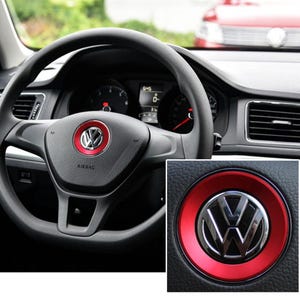 Può includere: Volante nero per auto con logo Volkswagen rosso e argento al centro. Il volante ha una trama simile alla pelle e il cruscotto è visibile sullo sfondo.