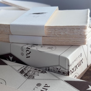 Könnte beinhalten: Ein Stapel cremefarbenes Papier mit Büttenrand, zusammengehalten von einer weißen Papierbanderole mit schwarzer Illustration. Darunter befindet sich eine weiße Schachtel mit schwarzen geometrischen Mustern, Gebäudezeichnungen und dem Text La Carta di Amalfi.
