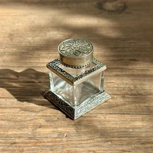 May include: A square glass inkwell featuring a decorative silver-toned metal base and a matching circular lid. The lid and the base are adorned with intricate embossed scrollwork patterns, while the clear glass body reveals a hollow interior.