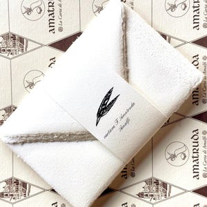 May include: A stack of off-white, textured handmade paper envelopes secured with a paper band. The band features a black ink illustration of a feather and the text "cartiera F. Amatruda Amalfi" printed in a serif font on the center.
