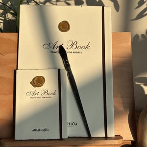 May include: Two cream-colored art books with dark brown elastic bands and gold wax seals on the covers. The text reads Art Book Travel Diary for Artists and amatruda. A black paintbrush with a silver ferrule rests diagonally across the books.